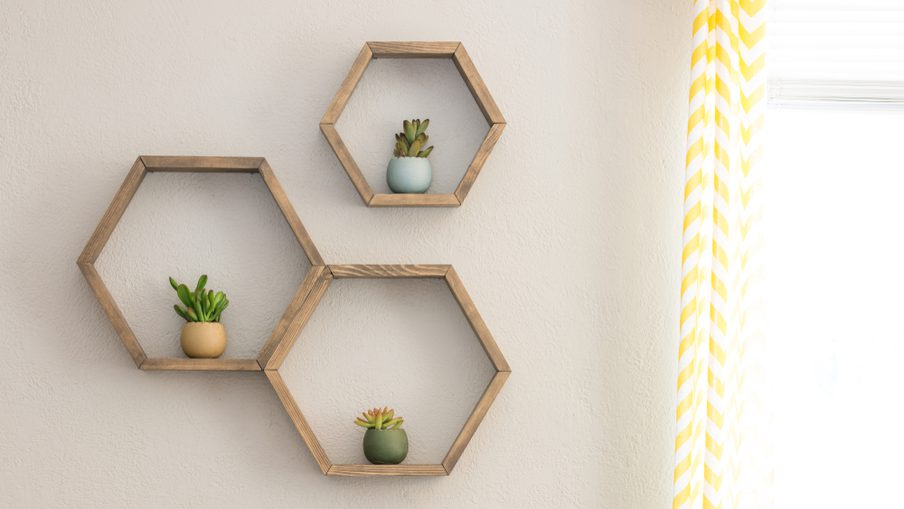 Three hexagonal wooden shelves on a beige wall, each holding a small succulent; bright window with a yellow-and-white striped curtain on the right.