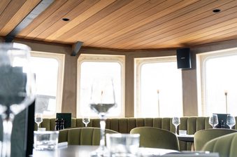 Dining area with a wood-paneled ceiling, olive-green booth seating, round tables set with wine glasses, and bright curved windows along the wall.