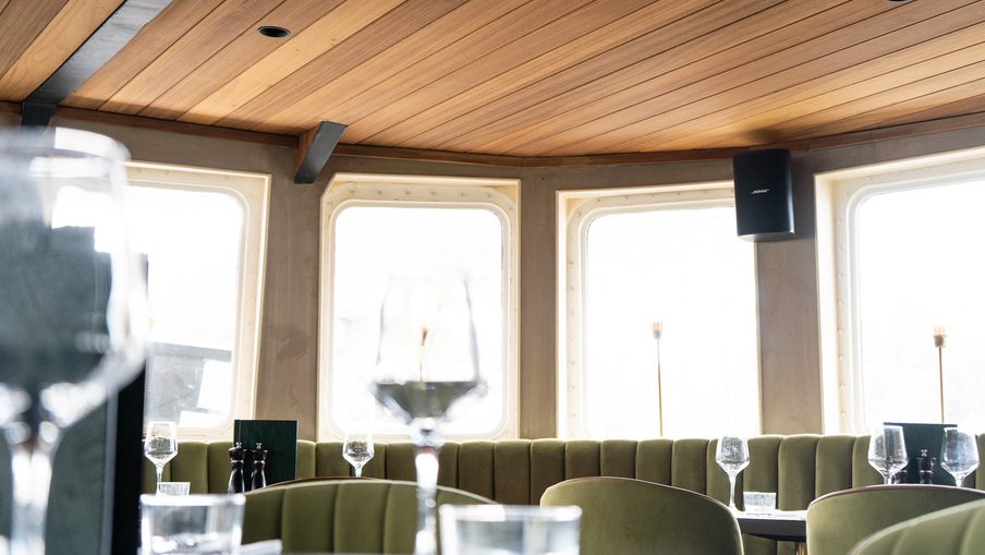 Dining area with a wood-paneled ceiling, olive-green booth seating, round tables set with wine glasses, and bright curved windows along the wall.