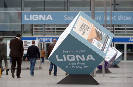 Large cube sculpture on a black base in a busy exhibition hall, displaying “LIGNA Next show 11-15 May 2015” with people walking in the background.