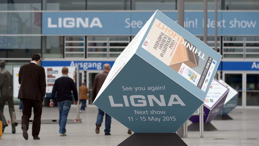Large cube sculpture on a black base in a busy exhibition hall, displaying “LIGNA Next show 11-15 May 2015” with people walking in the background.