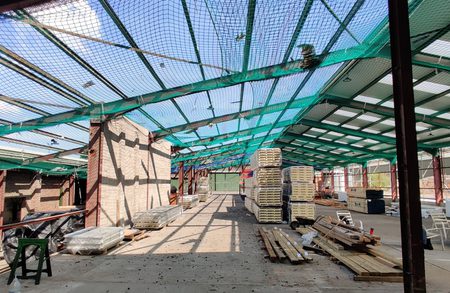 Indoor construction site with green safety netting and metal framework, brick wall on the left, stacks of pallets and lumber in the center, and a white van with chairs on the right.