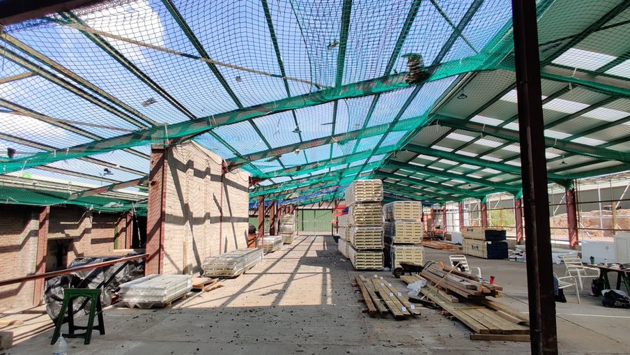 Indoor construction site with green safety netting and metal framework, brick wall on the left, stacks of pallets and lumber in the center, and a white van with chairs on the right.