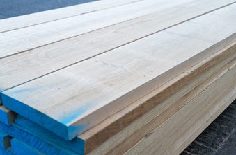 Stack of light wooden planks with blue-painted ends, showing wood grain, outdoors on a dark surface.