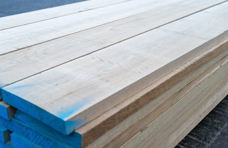 Stack of light wooden planks with blue-painted ends, showing wood grain, outdoors on a dark surface.