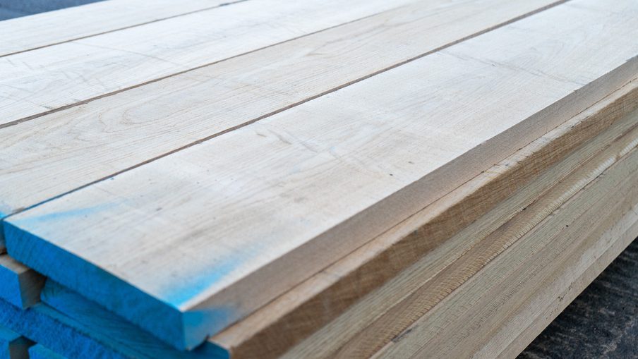 Stack of light wooden planks with blue-painted ends, showing wood grain, outdoors on a dark surface.