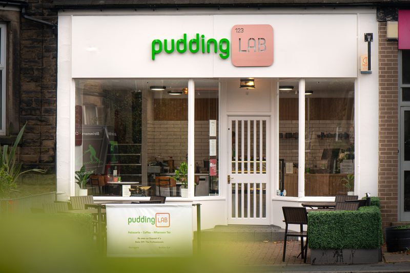 Storefront of pudding LAB café: white facade with a green “pudding” sign and pink LAB badge, large glass windows, white door, and outdoor seating with chairs and hedges.