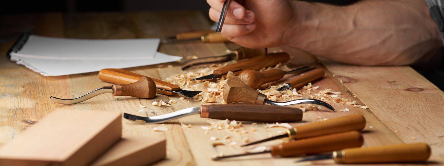 Close-up of a hand carving wood on a workbench, with gouges, chisels, wood shavings, and wooden blocks nearby.