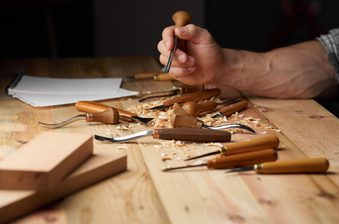 Close-up of a hand carving wood on a workbench, with gouges, chisels, wood shavings, and wooden blocks nearby.