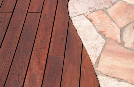 Curved edge where a reddish-brown wooden deck meets light pink stone paving; wood planks show grain, with a visible screw near the boundary.