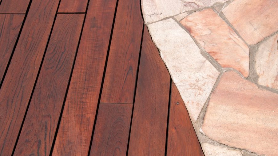 Curved edge where a reddish-brown wooden deck meets light pink stone paving; wood planks show grain, with a visible screw near the boundary.