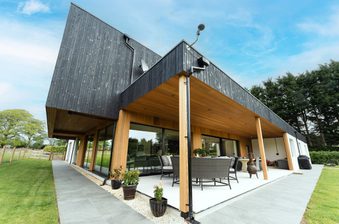 Modern house with a black slanted roof, timber columns, and glass walls; covered patio with wicker seating, potted plants, and a green lawn.