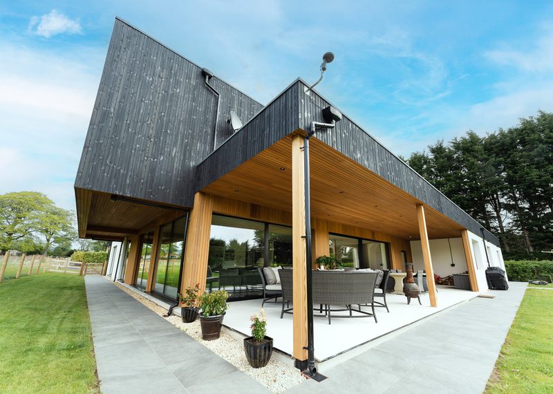 Modern house with a black slanted roof, timber columns, and glass walls; covered patio with wicker seating, potted plants, and a green lawn.