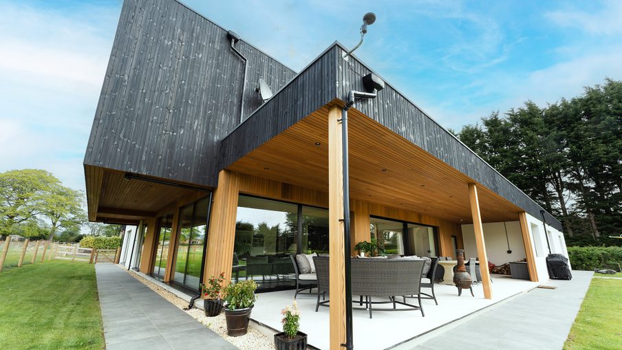 Modern house with a black slanted roof, timber columns, and glass walls; covered patio with wicker seating, potted plants, and a green lawn.