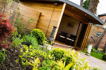 Wooden garden room with a curved roof and open veranda; two wicker chairs sit on a shaded porch, surrounded by lush shrubs and a brick path.