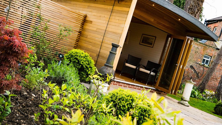 Wooden garden room with a curved roof and open veranda; two wicker chairs sit on a shaded porch, surrounded by lush shrubs and a brick path.
