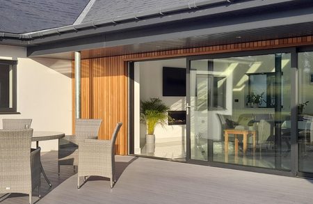 Modern house exterior with vertical wood cladding and large sliding glass doors opening to a bright living area; a grey deck with a wicker dining set on the patio.