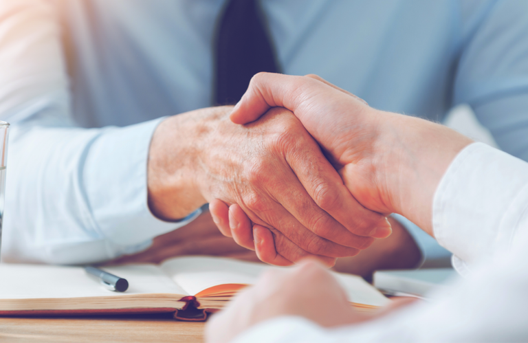 Two people shake hands across a desk with a glass of water, an open notebook, and a pen, suggesting a business meeting or agreement.