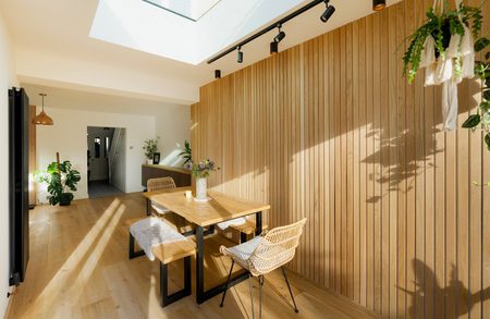 Bright dining area with a vertical wooden slat wall, skylight and track lighting; a wooden table with wicker chairs and a bench, potted plants, and sunlit shadows.