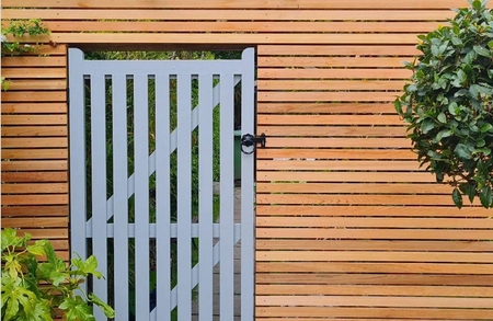 Blue-gray wooden gate set in a horizontal fence, with a black latch; green plants frame the sides and a garden path shows through behind the gate.