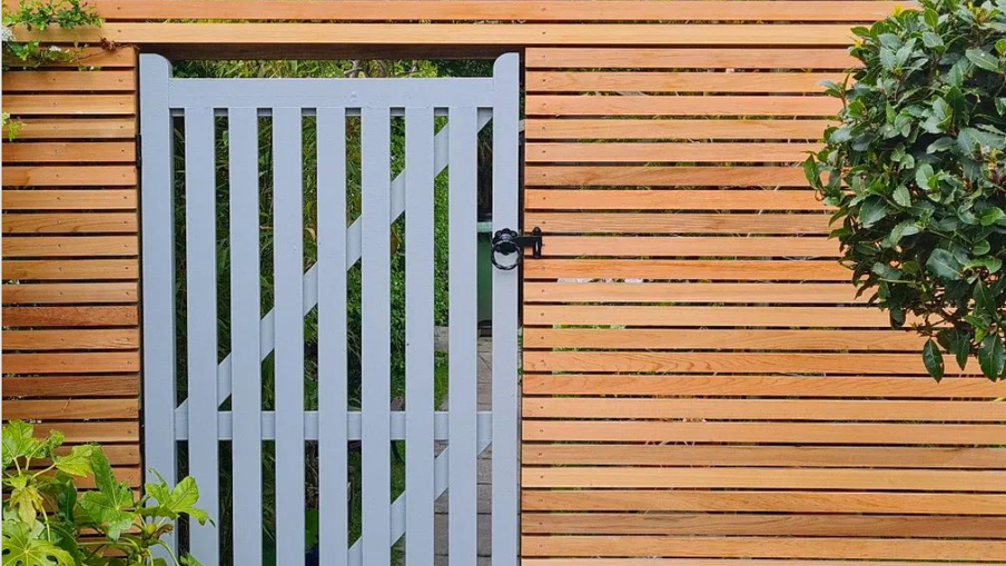 Blue-gray wooden gate set in a horizontal fence, with a black latch; green plants frame the sides and a garden path shows through behind the gate.