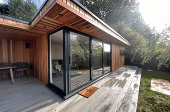 Modern wooden house with large black-framed sliding glass doors, a wide grey deck, and an outdoor dining area; star-pattern doormat and greenery around.