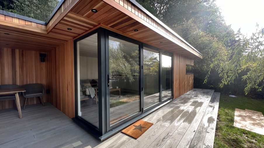Modern wooden house with large black-framed sliding glass doors, a wide grey deck, and an outdoor dining area; star-pattern doormat and greenery around.