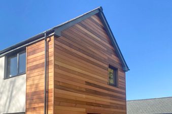 Two-story house with a wood-clad, horizontal plank facade on the right and white wall on the left, a small upper window, and a gutter downspout against a blue sky.