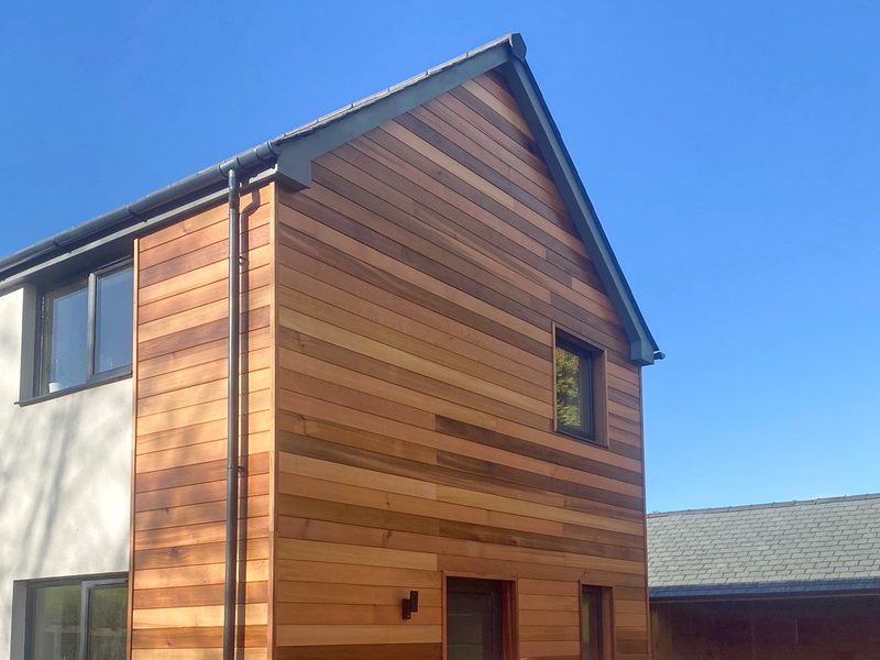 Two-story house with a wood-clad, horizontal plank facade on the right and white wall on the left, a small upper window, and a gutter downspout against a blue sky.