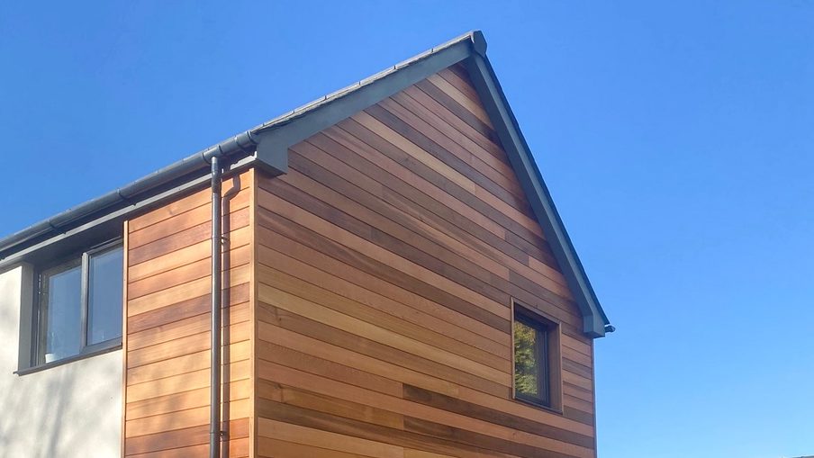 Two-story house with a wood-clad, horizontal plank facade on the right and white wall on the left, a small upper window, and a gutter downspout against a blue sky.