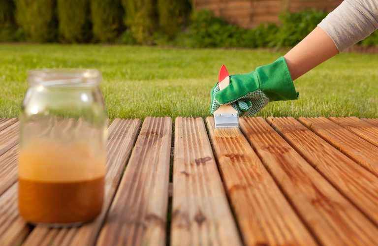 Hand in a green glove applies a small brush to stained wooden deck boards; a jar of orange wood stain sits on the left.