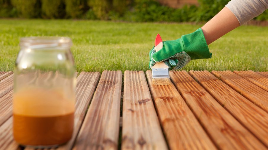 Hand in a green glove applies a small brush to stained wooden deck boards; a jar of orange wood stain sits on the left.