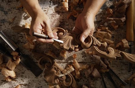 Hands carve an ornate wood scroll pattern with a chisel, surrounded by wood shavings on a workbench.