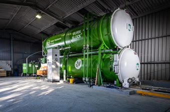 Inside a warehouse, a large green Burn Block industrial unit for timber treatments, with stacked cylindrical tanks, piping, and a control cabinet.