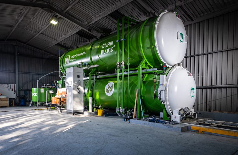 Inside a warehouse, a large green Burn Block industrial unit for timber treatments, with stacked cylindrical tanks, piping, and a control cabinet.