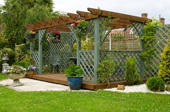Pergola with lattice posts and hanging flower baskets over a raised wooden deck, with potted plants and a gravel border framing a green lawn.