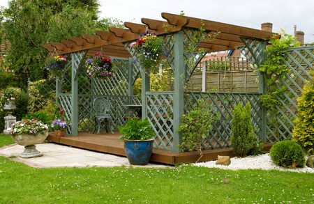 Pergola with lattice posts and hanging flower baskets over a raised wooden deck, with potted plants and a gravel border framing a green lawn.