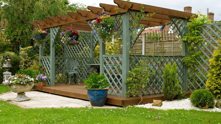 Pergola with lattice posts and hanging flower baskets over a raised wooden deck, with potted plants and a gravel border framing a green lawn.