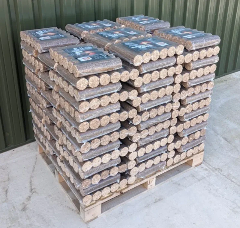 Stack of bagged wood pellets on a wooden pallet, outdoors beside a green corrugated metal wall.