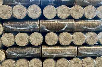 Stacks of cylindrical charcoal briquettes, dusty gray, tightly arranged in rows with some clear plastic wrap.