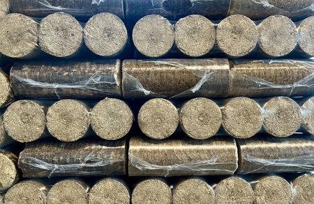 Stacks of cylindrical charcoal briquettes, dusty gray, tightly arranged in rows with some clear plastic wrap.