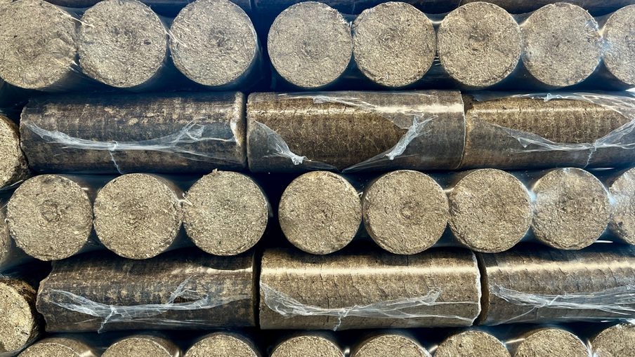 Stacks of cylindrical charcoal briquettes, dusty gray, tightly arranged in rows with some clear plastic wrap.