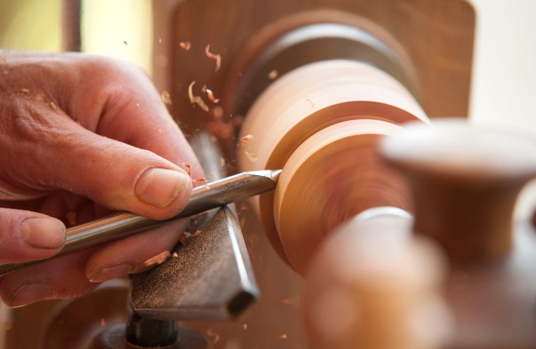 Close-up of hands shaping a spinning wooden piece on a lathe with a gouge, wood shavings flying as the tool cuts.