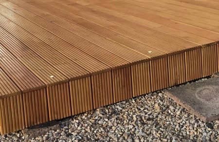 Warm brown wooden deck with narrow grooved boards and visible screws, raised above gravel with stone stepping pads nearby.