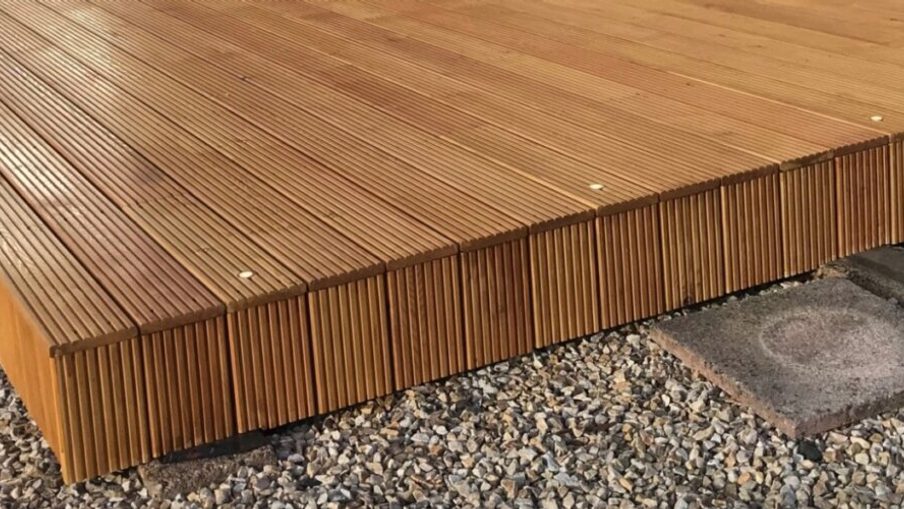 Warm brown wooden deck with narrow grooved boards and visible screws, raised above gravel with stone stepping pads nearby.