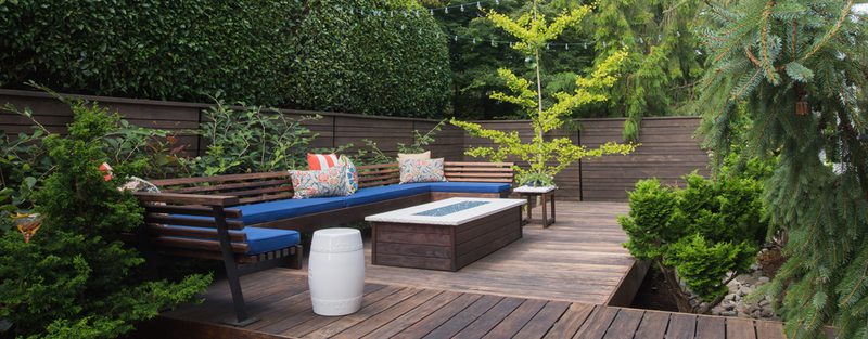 Cozy outdoor wooden deck with L-shaped bench, blue cushions and patterned pillows, a fire pit table, a white barrel stool, string lights, and greenery.