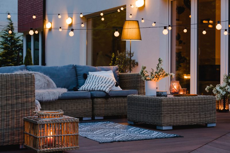Cozy outdoor lounge on a deck with a woven rattan sectional, striped pillow, string lights overhead, a wicker coffee table, and candles for warm ambiance.