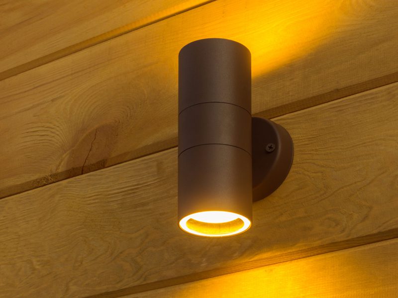 A dark cylindrical outdoor wall light mounted on wooden siding, casting warm yellow light downward.