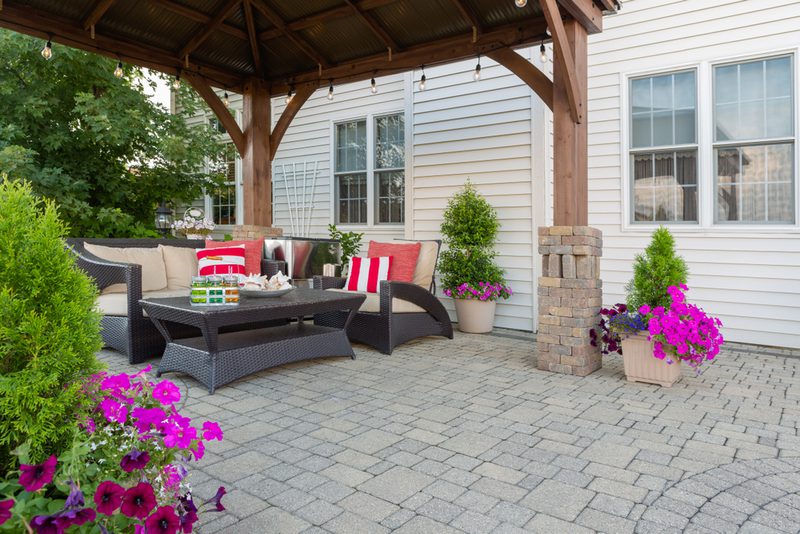 Covered patio with wicker sofa, cushions, red-striped pillows, glass coffee table, string lights, brick columns, and potted flowers beside a house.