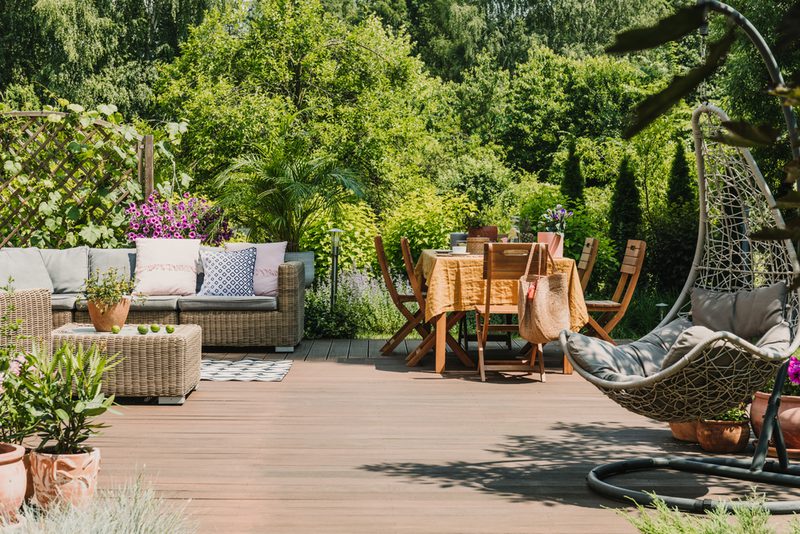 Sunlit backyard patio with a wicker sectional and colorful cushions on the left, a central wooden dining table with chairs, and a hanging chair on the right.